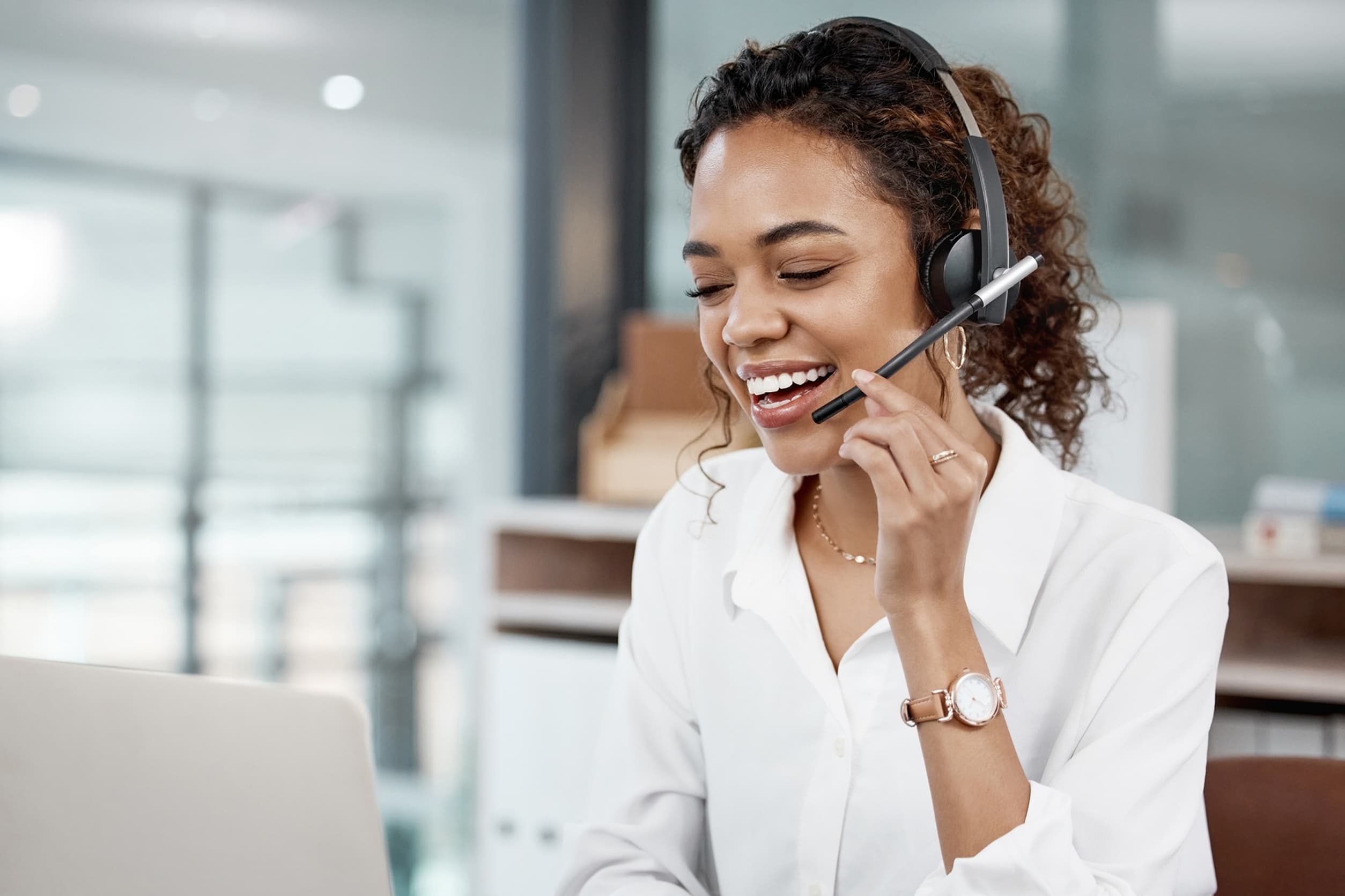 Happy young woman at her desk, speaking with someone through her headset in front of a computer