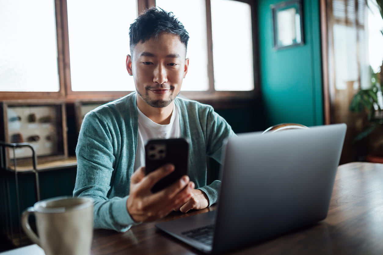 Young man sitting at a table with his laptop open, looking at his phone