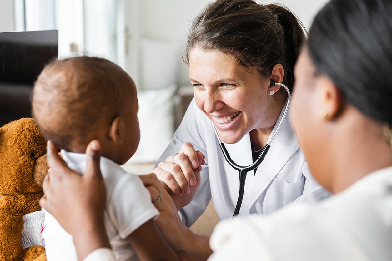 A smiling woman holds a baby while looking at a doctor in a bright, cheerful office setting