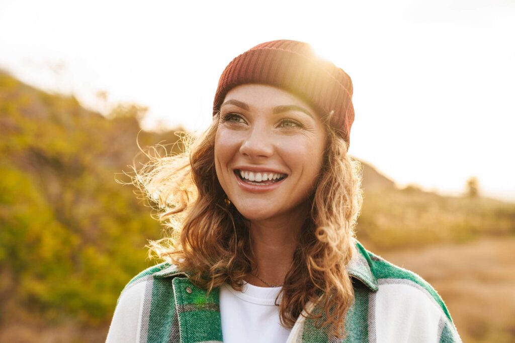 Young woman wearing hat and plaid shirt walking outdoors and smiling in the sunlight